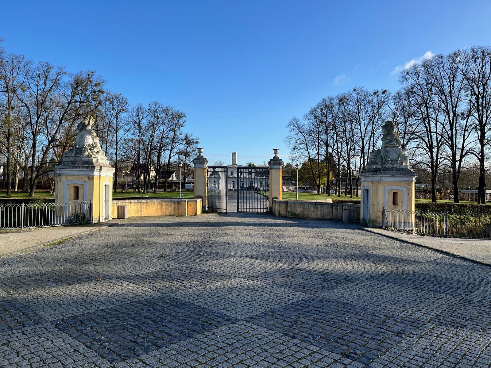 Blick durch ein schmiedeeisernes Tor auf einen gepflasterten Vorplatz mit zwei gelben Torhäusern, die von steinernen Skulpturen gekrönt sind.