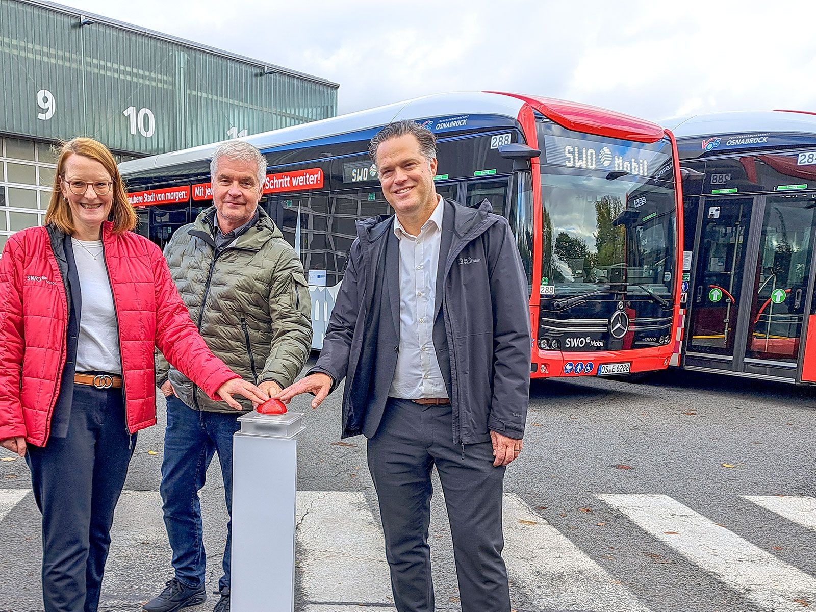 Eine Frau und zwei Männer drücken auf einen roten Knopf und stehen dabei vor einem Bus.