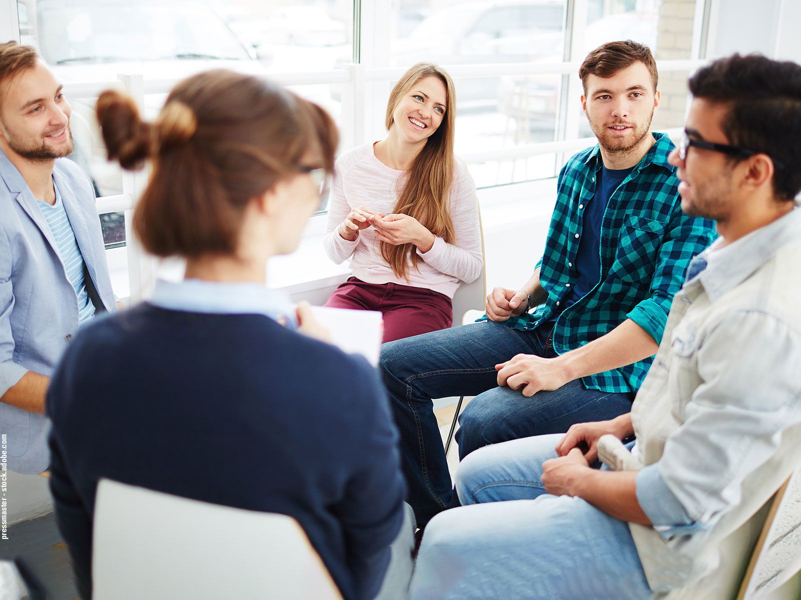 A group of young adults is sitting together in an informal discussion in a bright room, engaging in lively conversation.