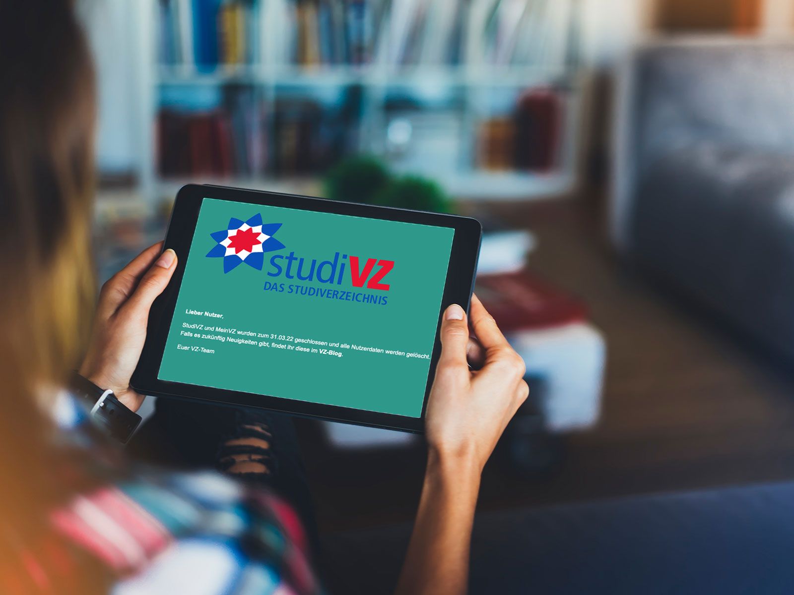 Eine Person hält ein Tablet, auf dem die Abschiedsmeldung der Plattform "studiVZ" mit dem Logo und dem Hinweis auf die Schließung zum 31.03.2022 vor einem unscharfen Wohnzimmerhintergrund zu sehen ist.