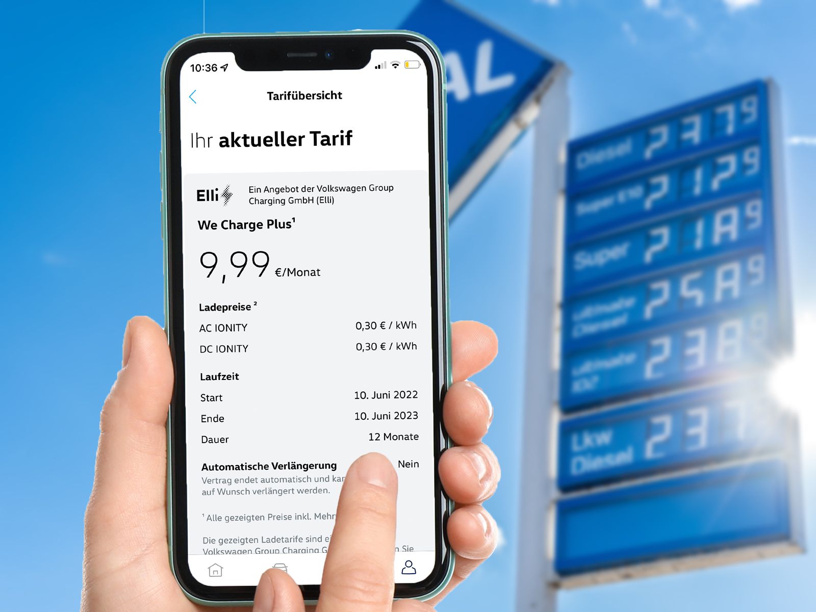 A smartphone shows the app interface of an electric vehicle charging tariff from the Elli brand, displaying pricing information, while in the blurred background a blue fuel station price sign shows high fuel prices for diesel and petrol (Super)