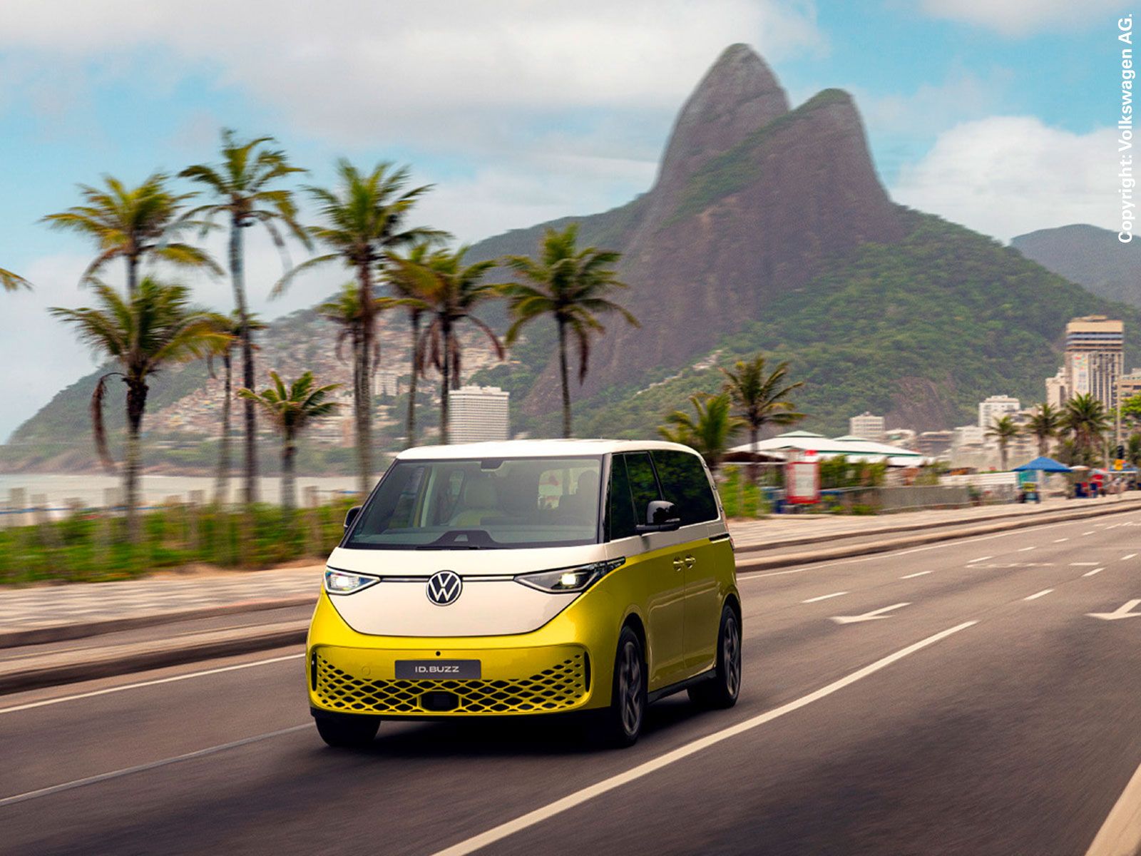 A yellow-and-white VW ID. Buzz is driving along a multi-lane coastal road in Rio de Janeiro, passing palm trees, with the iconic Two Brothers mountain (Morro Dois Irmãos) in the background.