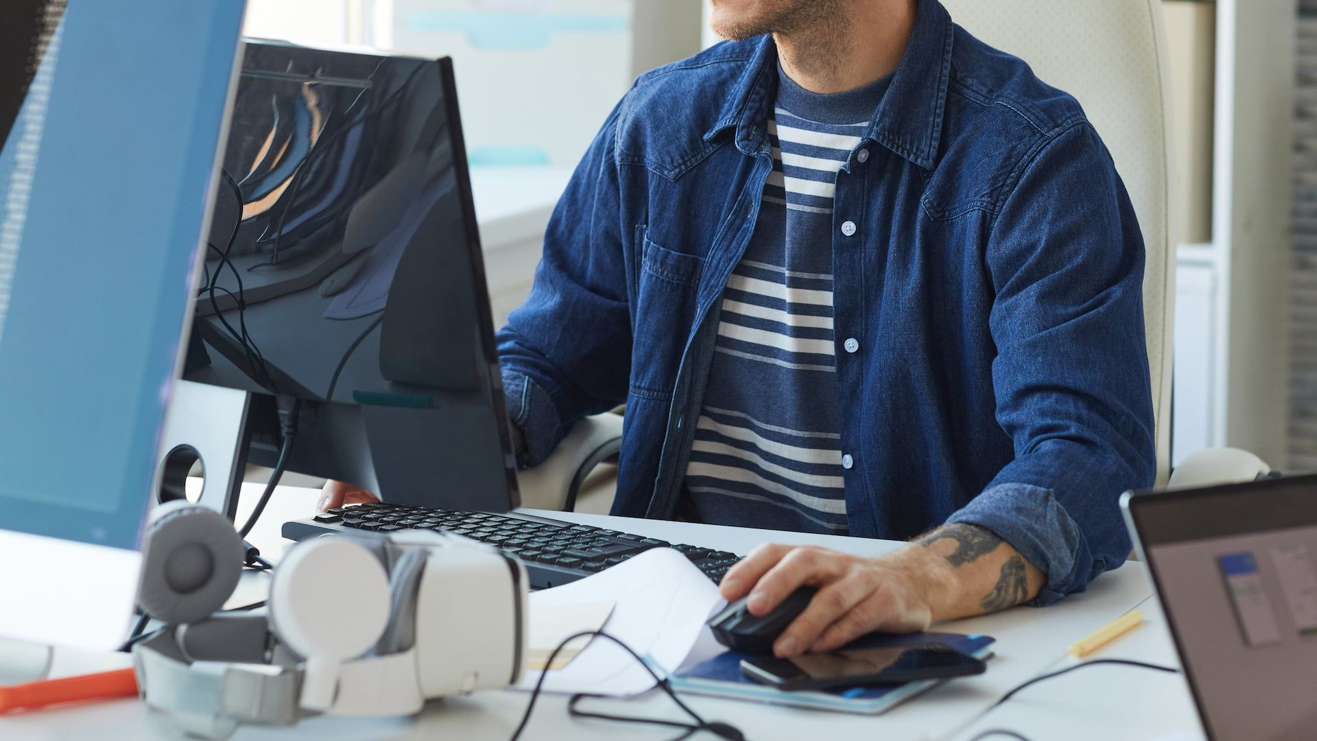 A man in a blue denim shirt is sitting at a desk and working on his computer.