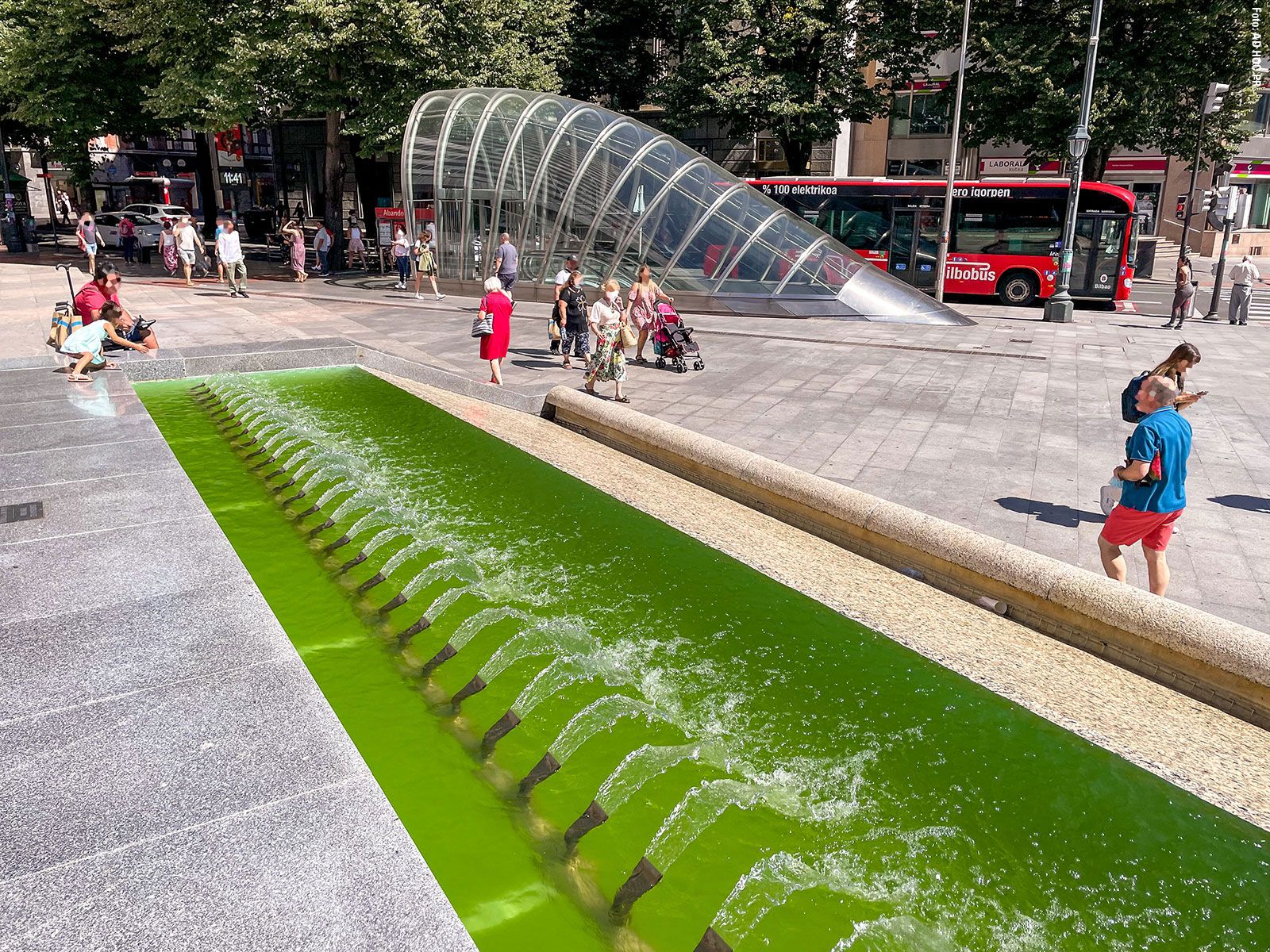 A modern glass subway entrance with a curved design on a busy city square, in the foreground a long rectangular fountain basin with greenish shimmering water and numerous small jets of water.