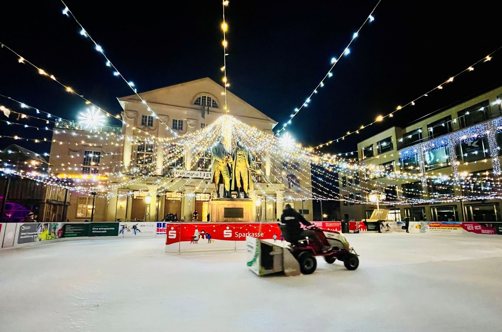 Night scene in Weimar with an artificial ice rink in the foreground, with a Zamboni ice resurfacing machine driving across it, in front of the illuminated Goethe-Schiller Monument and the Deutsches Nationaltheater, under a sky filled with decorative lights.